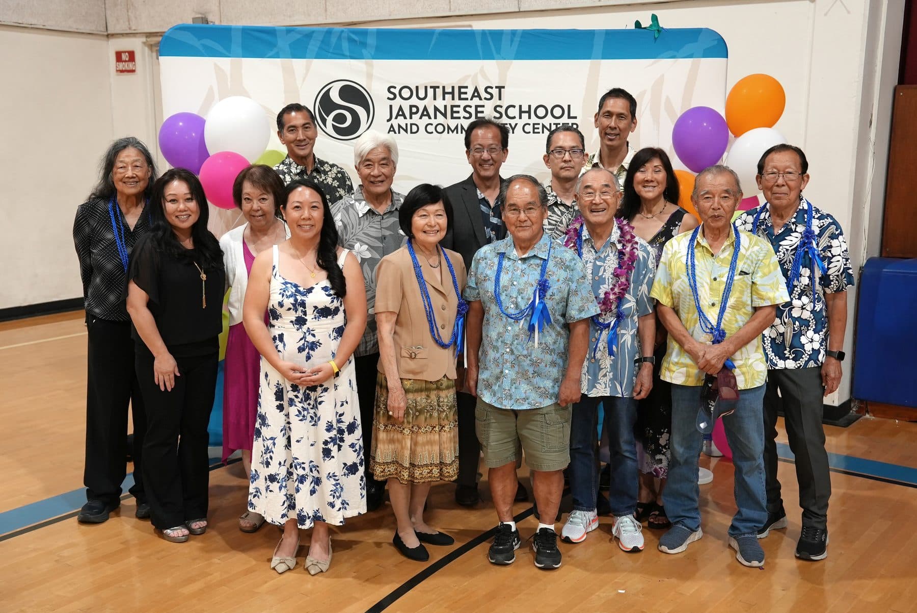 The 2025 board of directors standing together in the gym in front of the center's banner, many wearing blue leis