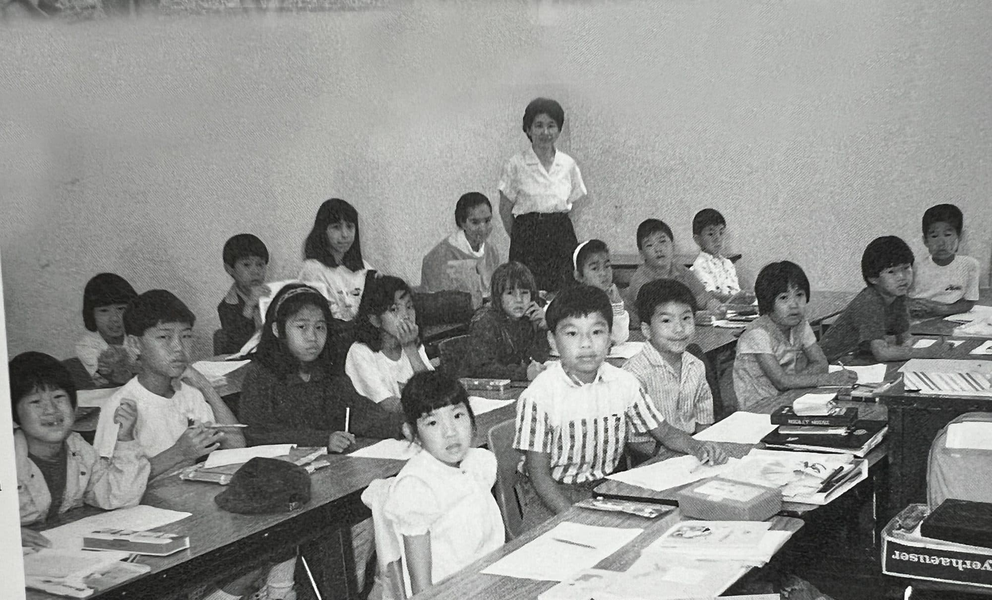 A black-and-white photo of a teacher standing before rows of students seated at desks in a Japanese-language classroom