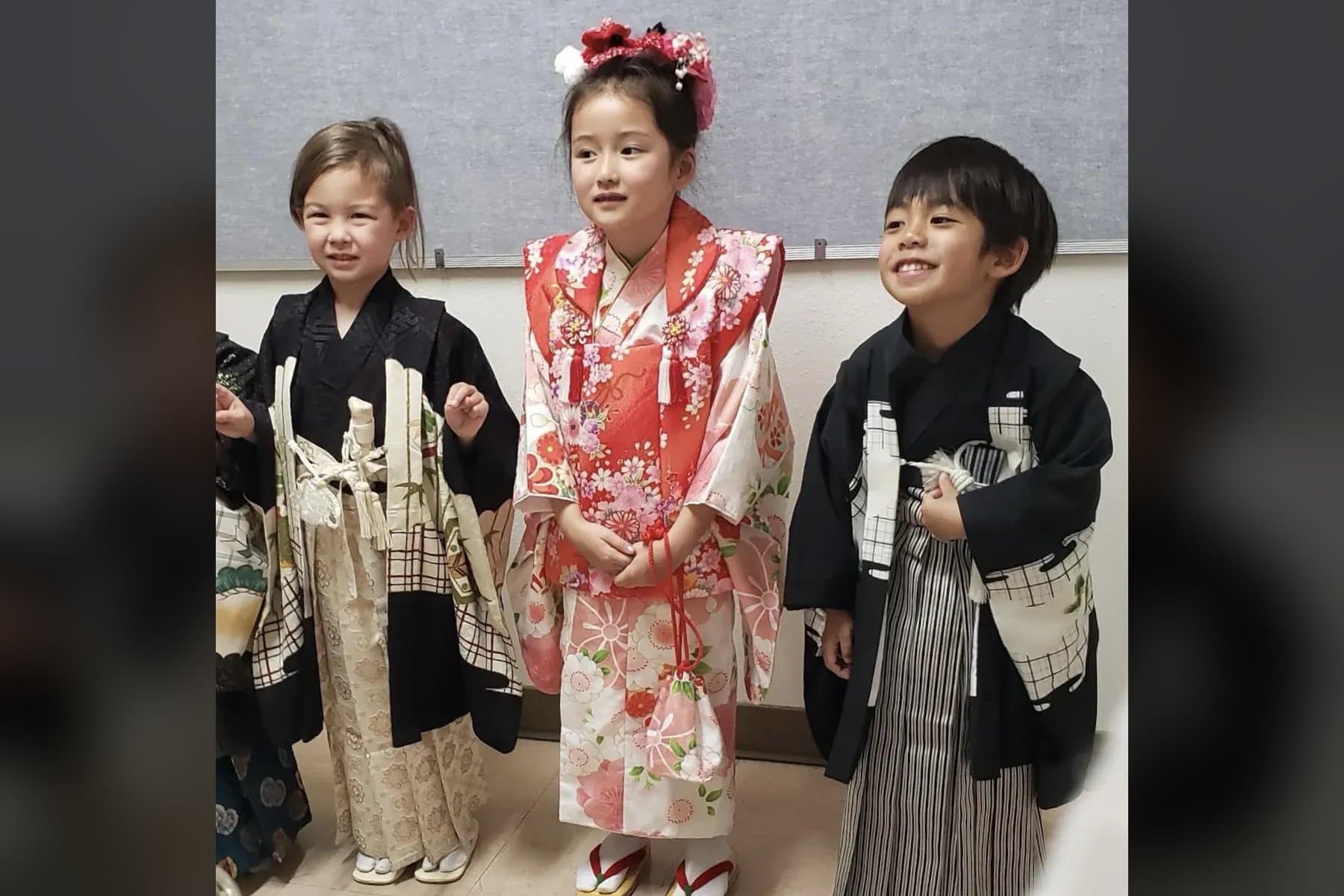 Three young students dressed for Shichi-go-san: a girl in a red and pink floral kimono with flowers in her hair, between two smiling boys in black haori and hakama